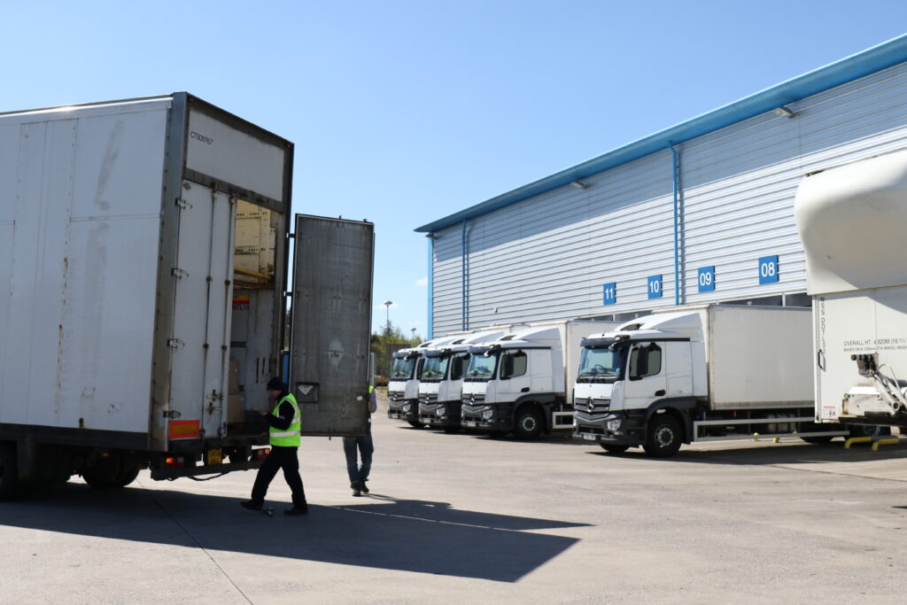 A national NHS Scotland distribution and logistics centre with several delivery lorries parked outside loading bays. A worker in high-visibility clothing stands beside an open trailer while goods are being loaded or unloaded. The image represents supply chain operations, warehousing and the transportation of goods and services.