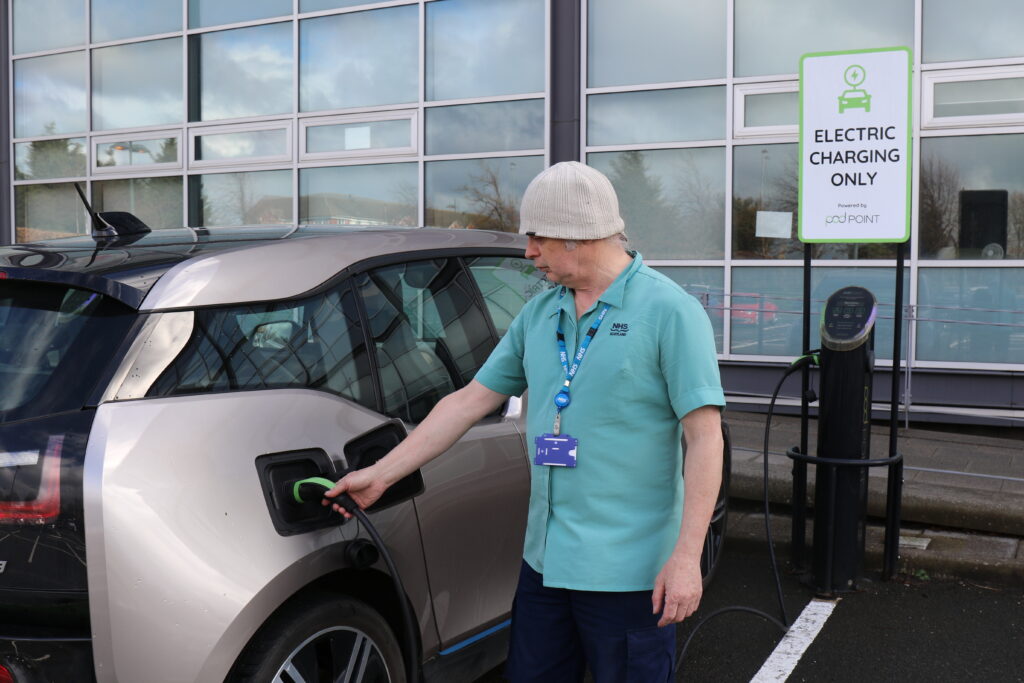 A member of staff connects an electric vehicle to a charging point outside a modern healthcare building. A sign beside the charging station indicates that the parking space is reserved for electric vehicle charging. The image highlights the adoption of low-emission transport, electric vehicle infrastructure and sustainable travel within healthcare settings.