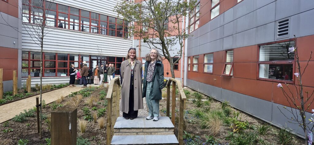 People stand and walk within a landscaped courtyard between modern healthcare buildings. Raised planting beds, young trees and accessible pathways create a green outdoor space within the hospital environment. The image highlights the integration of biodiversity, greenspace and wellbeing-focused design within healthcare estates.