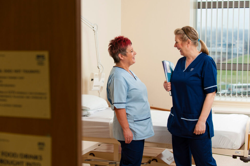 Two healthcare staff members stand and talk beside a hospital bed in a patient care room. One person holds a folder while they discuss information in the clinical environment. Natural light enters through large windows fitted with blinds. The image highlights teamwork, communication and collaboration in the delivery of patient care.