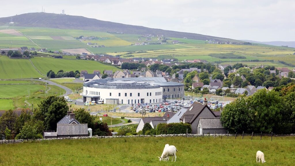 The Balfour hospital healthcare facility is situated within a rural landscape, surrounded by green fields, rolling hills and a nearby village. The building overlooks open countryside, with farmland visible in the foreground and hills in the distance. The image highlights the connection between healthcare services, local communities and the natural environment in a rural setting.